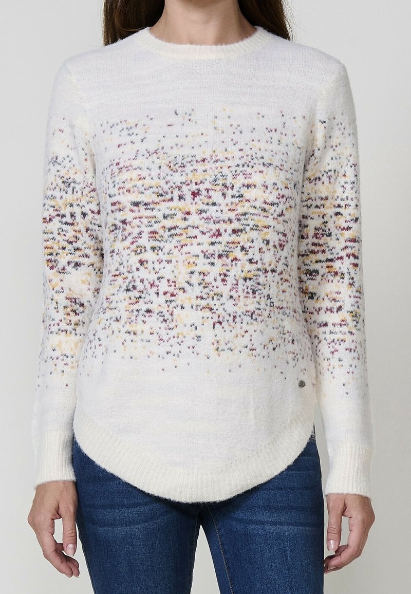 Femme portant un pull en maille blanc avec un motif tacheté multicolore et un jean bleu, debout devant un fond gris uni.