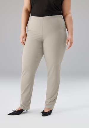 Kurvige Person, die beige eng anliegende Hosen, ein schwarzes ärmelloses Oberteil und schwarze spitz zulaufende Schuhe mit niedrigem Absatz trägt und vor einem neutralen Hintergrund steht.