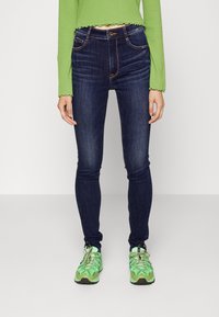 Mörkblå skinny jeans med framfickor, guldtrådstickning och hög midja, matchade med en ljusgrön långärmad crop top och gröna sneakers.