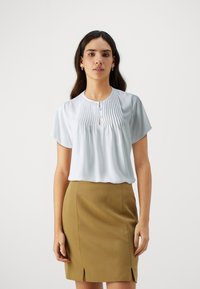 Blusa azzurra a maniche corte con pieghe verticali, scollatura a chiave e dettagli con bottoni, abbinata a una gonna aderente color khaki con spacchi.
