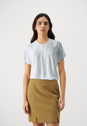Blusa azzurra a maniche corte con pieghe verticali, scollatura a chiave e dettagli con bottoni, abbinata a una gonna aderente color khaki con spacchi.