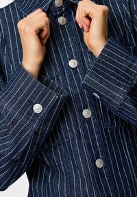 Chaqueta de denim con rayas verticales blancas, que presenta un cuello alto, botones a presión plateados y bolsillos con solapa. Tejido azul con textura suave.