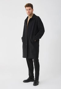 Manteau long en mélange de laine noire avec une coupe ajustée, design à simple boutonnage, grands boutons et poches latérales. Associé à un pantalon noir slim.