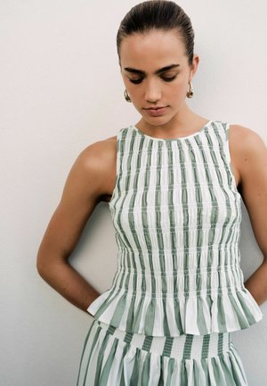 Woman with dark hair pulled back wears sleeveless white top with green vertical stripes and ruffled hem, looking downward against plain wall.