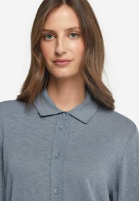 Ljust blå skjorta med krage, tillverkad av texturerad tyg. Har en knäppning med tre knappar och något rullade ärmar.