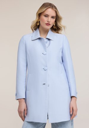 Kobieta w jasnoniebieskim płaszczu zapinanym na guziki z długimi rękawami, do tego teksturowane jasnoniebieskie spodnie i złote kolczyki.