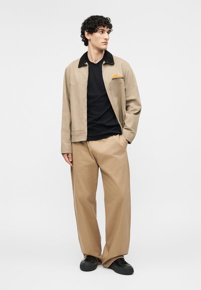 Νεαρός άνδρας στέκεται, φορώντας μπεζ μπουφάν με μαύρο γιακά, μαύρο πουκάμισο, φαρδιά μπεζ παντελόνια και μαύρα παπούτσια, σε απλό ανοιχτό φόντο.