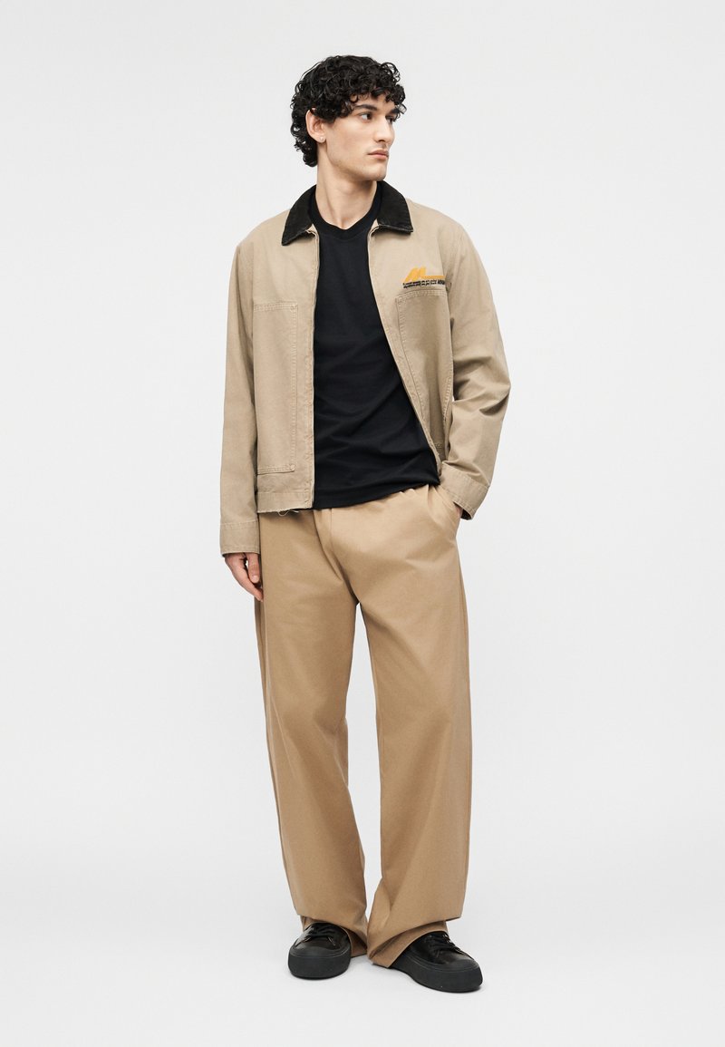 Jonge man staand, draagt een beige jas met zwarte kraag, zwart overhemd, wijde beige broek en zwarte schoenen, voor een effen lichte achtergrond.