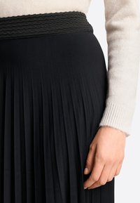 Falda plisada negra con cintura fruncida, combinada con un suéter beige claro que muestra puños acanalados. Vista en primer plano, centrada en la textura y los detalles.
