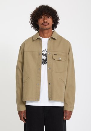 Mann mit lockigem Haar, der eine beige Jacke mit Knöpfen über einem weißen T-Shirt mit Grafik und schwarzen Hosen trägt, vor einem schlichten Hintergrund steht.