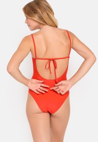Maillot de bain une pièce rouge avec des bretelles fines, un dos profond et une fermeture à nouer. Texture lisse sans motifs.