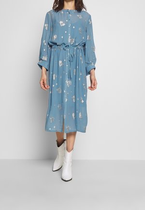 Woman wearing a blue midi dress with silver floral patterns and white cowboy boots, standing against a plain light gray background.