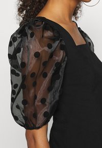 Czarna sukienka z puchatymi, przezroczystymi rękawami w duże czarne groszki. Strukturalny gorset z kwadratowym dekoltem i teksturowaną tkaniną.