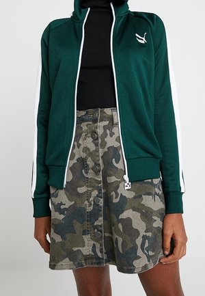 Femme portant une veste zippée Puma vert foncé, un top noir et une jupe camouflage à boutons sur le devant.
