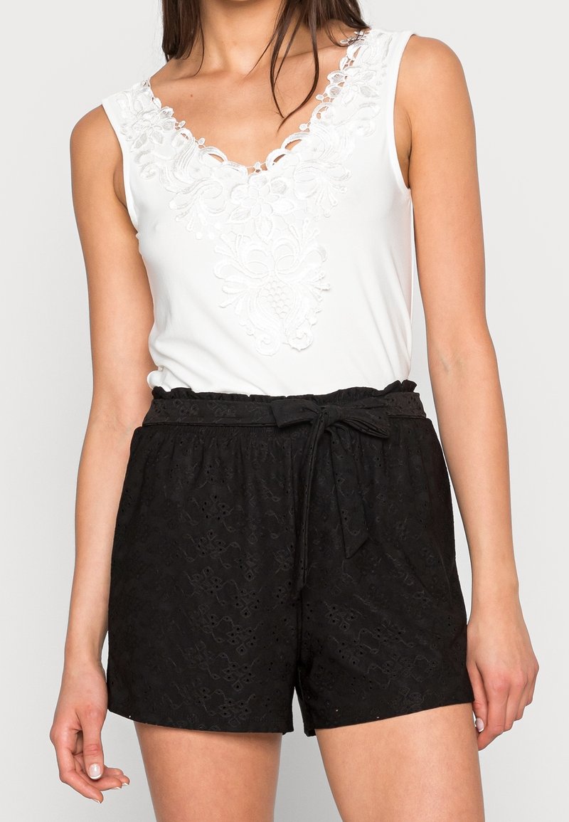 Femme portant un haut blanc sans manches avec un détail en dentelle florale et un short noir taille haute avec une ceinture nouée et un motif ajouré subtil.