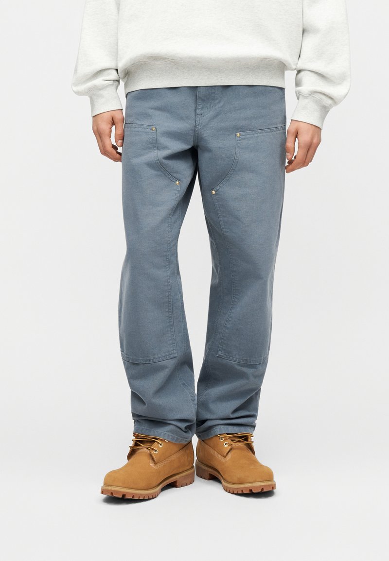 Man wearing blue work pants with front pockets and tan work boots, standing against a plain light background.