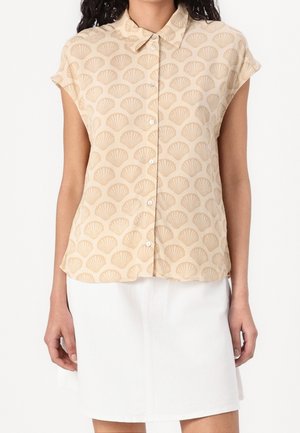 Beige overhemd met korte mouwen en knopen met schelpenpatroon, gecombineerd met een witte knielange rok bij een staand persoon.