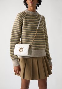 Προσωπικός λευκός τσάντα με ραφή, με χρυσή λεπτομέρεια, φορεμένη χιαστί. Διαθέτει αλυσίδα και ορθογώνιο σχήμα. Είναι επίσης ορατό ένα ρίγες πουλόβερ και φούστα με πτυχώσεις.