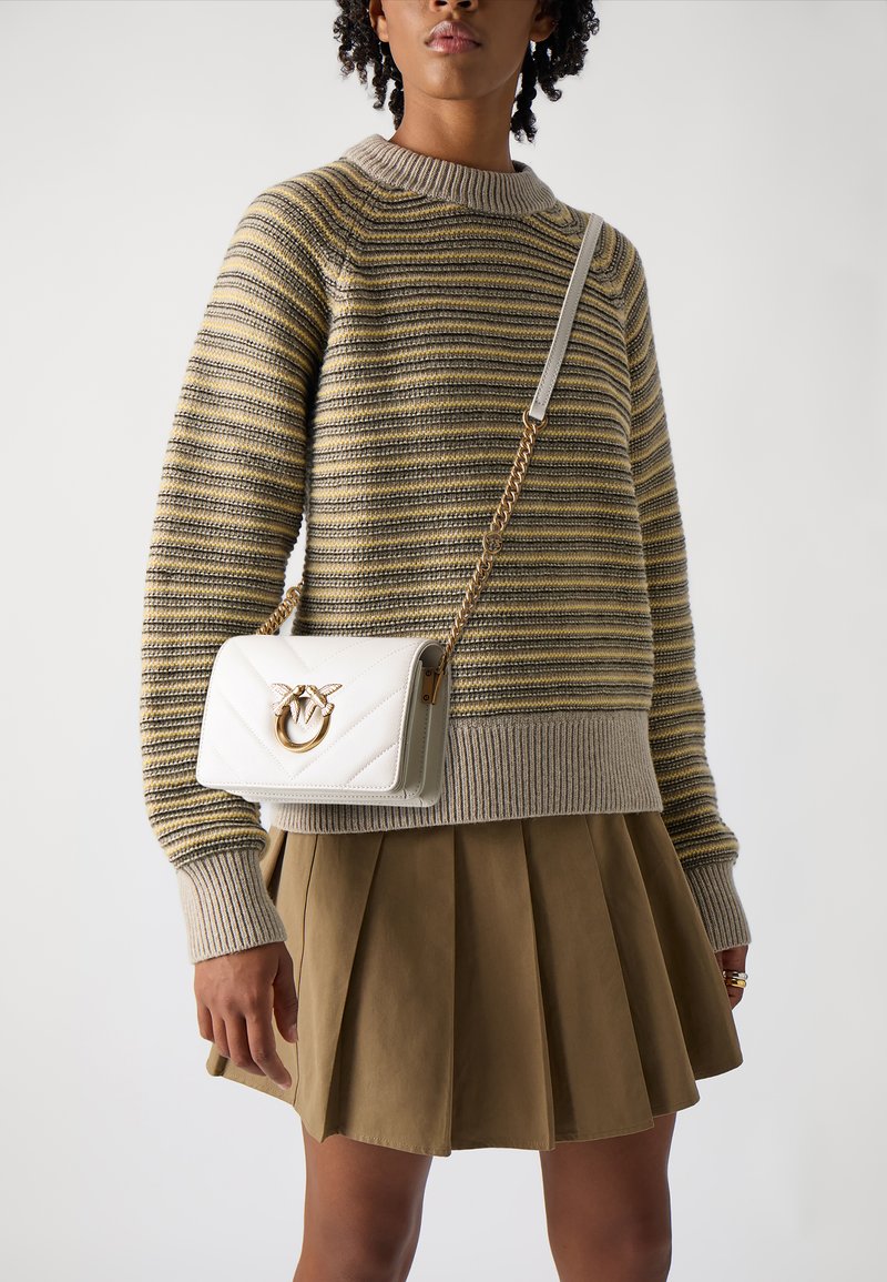 Προσωπικός λευκός τσάντα με ραφή, με χρυσή λεπτομέρεια, φορεμένη χιαστί. Διαθέτει αλυσίδα και ορθογώνιο σχήμα. Είναι επίσης ορατό ένα ρίγες πουλόβερ και φούστα με πτυχώσεις.