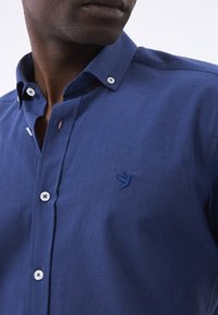 Camisa de botón azul marino con cuello regular, que cuenta con botones blancos y un logo de pájaro bordado en el lado izquierdo del pecho. Textura suave.