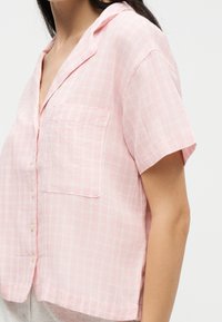 Femme portant une chemise rose à manches courtes à carreaux avec une poche poitrine et un col ouvert, montrée des épaules jusqu'à la taille.