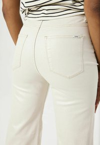 Beige high-waisted jeans met een glanzende textuur, twee achterzakken en contrasterende stiksels. Gecombineerd met een zwart-wit gestreept shirt.