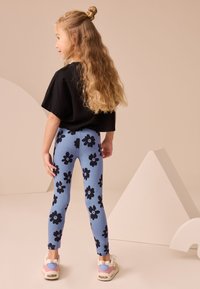 Kék leggings fekete virágmintákkal, fekete rövid ujjú felsővel és színes sportcipőkkel, hátulnézetből.