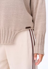 Beżowy sweter z dzianiny z prążkowanym dołem i mankietami, z małą metką po boku. W zestawieniu z jasnymi spodniami w paski.