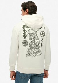 Hombre que lleva una sudadera blanca con un tigre negro, bambú y símbolos de estilo japonés impresos en la parte posterior, combinada con jeans azules.