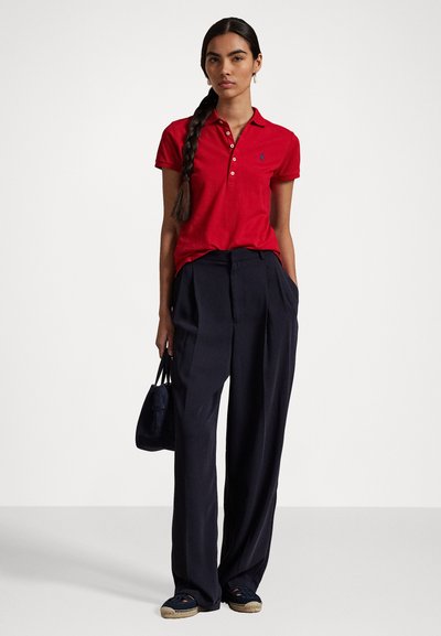 Red polo shirt with a collar, buttons, and logo, paired with dark blue wide-leg trousers. Model holds a small black bag, wearing navy espadrilles.