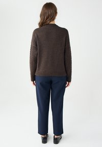 Brązowy sweter dzianinowy z detalami w warkocze na rękawach, zestawiony z granatowymi spodniami i czarnymi butami. Neutralne tło bez dodatkowych elementów.