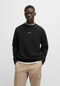 Svart sweatshirt med rund hals och ribbade muddar, med "BOSS"-logotyp i vitt. Bärs över en vit skjorta, ihop med khaki cargobyxor.