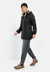 Zwarte pufferjack met horizontaal gestikte design, ritsluiting aan de voorkant en leren accenten. Gecombineerd met grijze jeans en bruine enkellaarsjes.