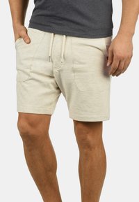 Bermudas beige para hombre hechas de tejido suave, con bolsillos laterales, cintura con cordón y un ajuste relajado. Textura lisa con un minimalismo en los detalles.