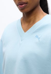 Κοντινό πλάνο ενός ατόμου που φοράει γαλάζιο φούτερ με V-λαιμόκοψη και μικρό κεντημένο λογότυπο Puma στο στήθος.