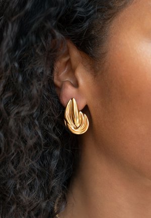 Pendientes color dorado con un diseño ondulado y escultórico que presenta costillas texturizadas y un acabado suave, sujetos a una oreja perforada.