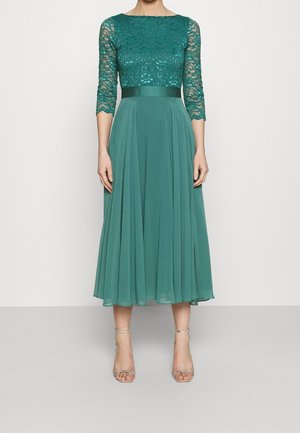 Robe midi bleu sarcelle avec manches trois-quarts en dentelle, taille ajustée, jupe fluide, associée à des sandales à talons hauts argentées avec bride à la cheville.