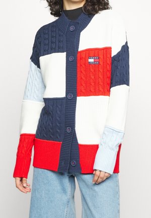 Persoon draagt een gebreid vest met knopen in kleurvlakken van marineblauw, rood, wit en lichtblauw, met een Tommy Jeans-logo op de borst.