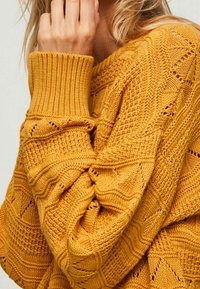 Gestrickter Pullover in leuchtendem Senfgelb mit strukturierten Mustern, gerippten Bündchen und lockerer Passform. Verziert mit offenen Strickakzenten.