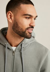 Grauer Zip-Up-Hoodie aus weichem Stoff, mit einer Kapuze mit Kordelzug und silbernen Hardware-Details. Das Model zeigt eine Nahaufnahme des Kragens und des Gesichts.
