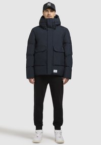 Marineblå pufferjakke med hætte, der har to forlommer, lynlåslukning og et logo mærke. Sammen med sorte sweatpants og hvide sneakers.