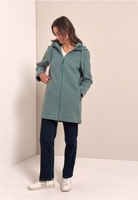 Turkoosi fleece-takki, jossa on huppu, täydellinen vetoketju ja sivutaskut, yhdistettynä tummansinihin housuihin ja valkoisiin lenkkareihin neutraalilla taustalla.