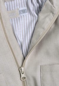 Lichtbeige, ritsjack met een textuur aan de buitenkant en een wit en bruin gestreepte binnenvoering. Inclusief een klein pocketdetail.