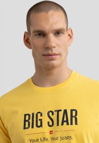 Dzeltena kokvilnas T-krekls ar tekstu "BIG STAR" treknrakstā zilā krāsā, ar drukātu saukli zem tā. Īsas piedurknes un apaļa kakla izgriezums.