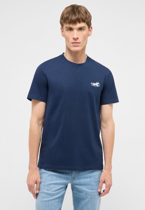Junger Mann trägt ein dunkelblaues kurzärmeliges T-Shirt mit kleinem weißem Pferdelogo auf der linken Brust, kombiniert mit hellblauen Jeans.
