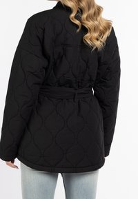 Svart vadderad jacka med bälte i midjan, mjuk textur, oversize-passform och puffad design med rund krage och diskret sömnadsdetalj.
