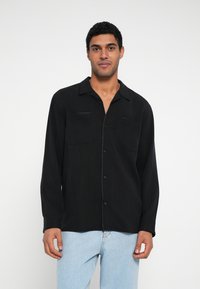 Musta pitkähihainen paita, jossa on napillinen edusta, kaksi rintataskua ja kaulus. Valmistettu kevyestä kankaasta, yhdistetty vaaleansiniin farkkuihin.