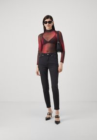 Gjennomsiktig langermet topp med røde og svarte geometriske mønstre, kombinert med tettsittende svarte jeans og svarte høye hæler med sirkulære detaljer.