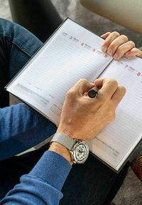 Une montre en mailles argentées orne une main tenant un stylo noir au-dessus d'un carnet blanc rempli de pages lignées et de marqueurs de dates.