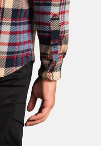 Koszula flanelowa w kratę z wzorem w odcieniach beżu, czerwieni i niebieskiego; czarne guziki; mankiety z podwinięciem; w zestawie z czarnymi spodniami.